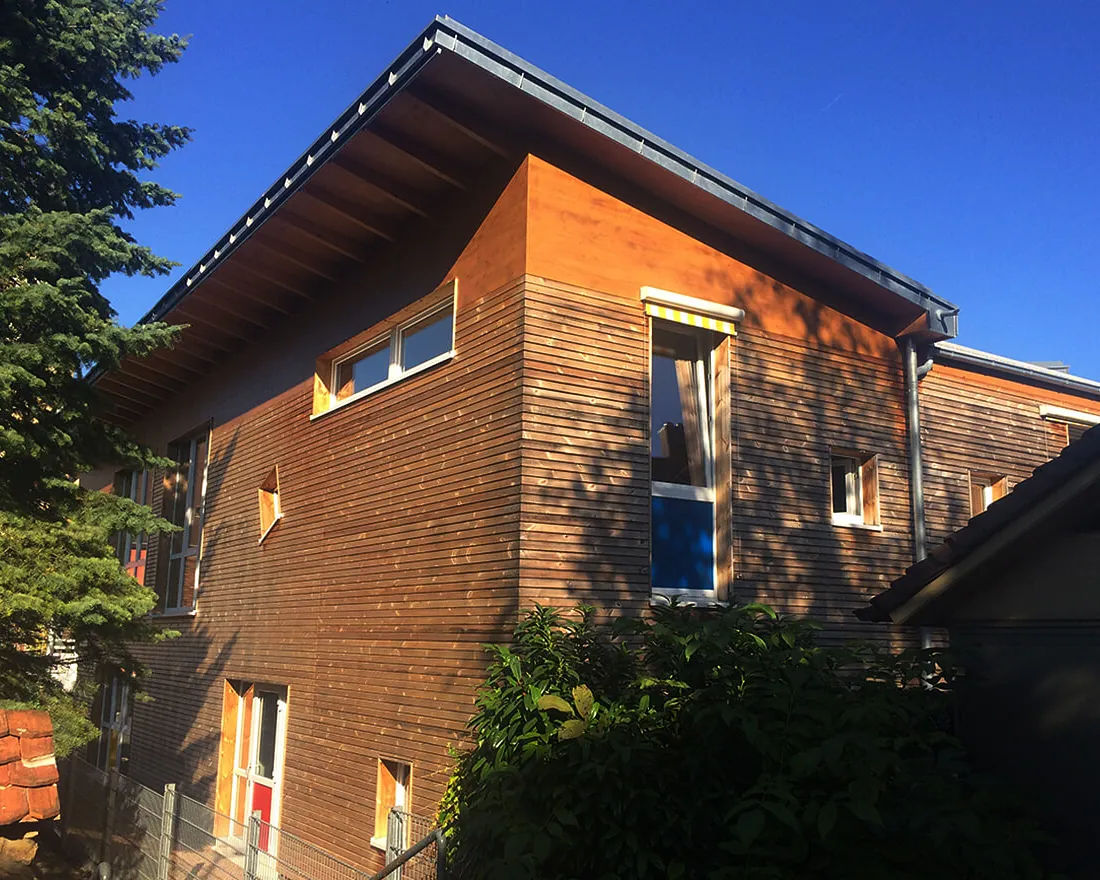 Modernes zweistöckiges Gebäude aus Holz mit großen Fenstern, umgeben von Bäumen und Sträuchern