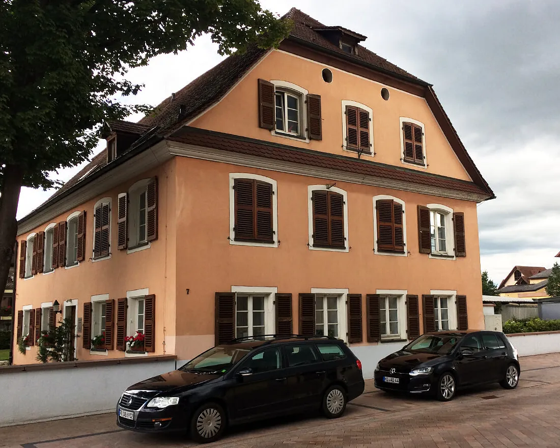 Zweistöckiges historisches Wohnhaus mit Satteldach, hellorange Fassade und braunen Fensterläden, zwei geparkte Autos davor