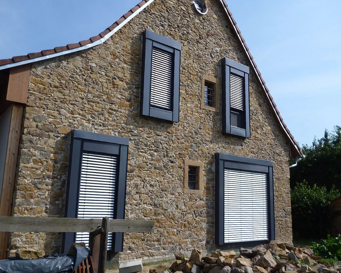 Steinernes Haus mit drei Fenstern und weißen Rollläden, darunter Holzstapel und blauer Himmel