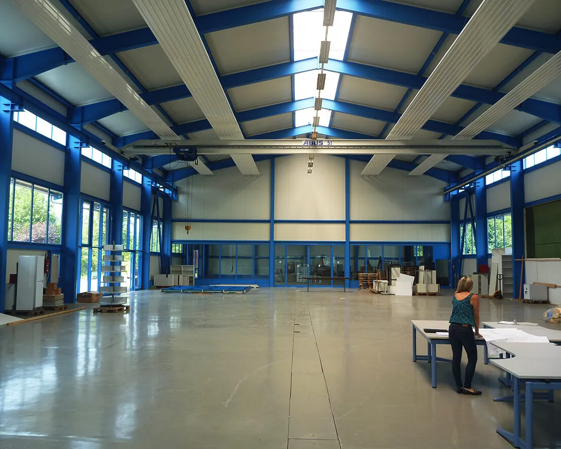 Große, helle Industriehalle mit blauen Stahlträgern und Oberlichtern, eine Person steht an einem Tisch mit Plänen