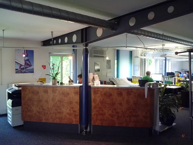 Büro mit Empfangstresen aus Holz, zwei Personen an Schreibtischen, große Fenster und sichtbare Lüftungsrohre an der Decke