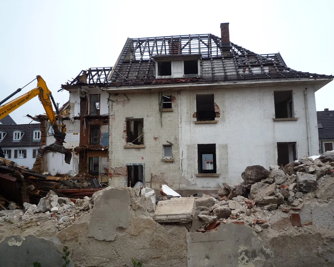 Abbruch eines zweistöckigen Hauses mit teilweise entferntem Dach und Bagger, Trümmer im Vordergrund