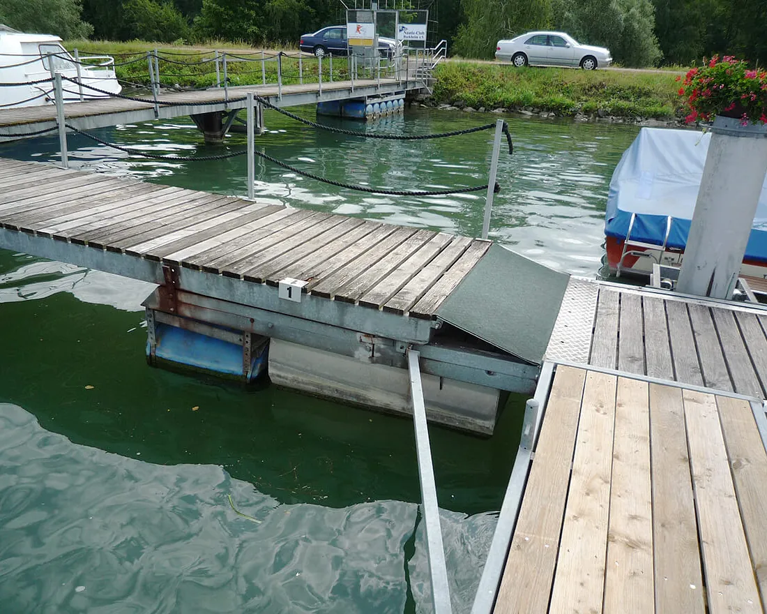 Holzsteg mit Metallgeländer über einem Gewässer, daneben ein Boot mit blauem Schutzcover und im Hintergrund Autos und Grünfläche
