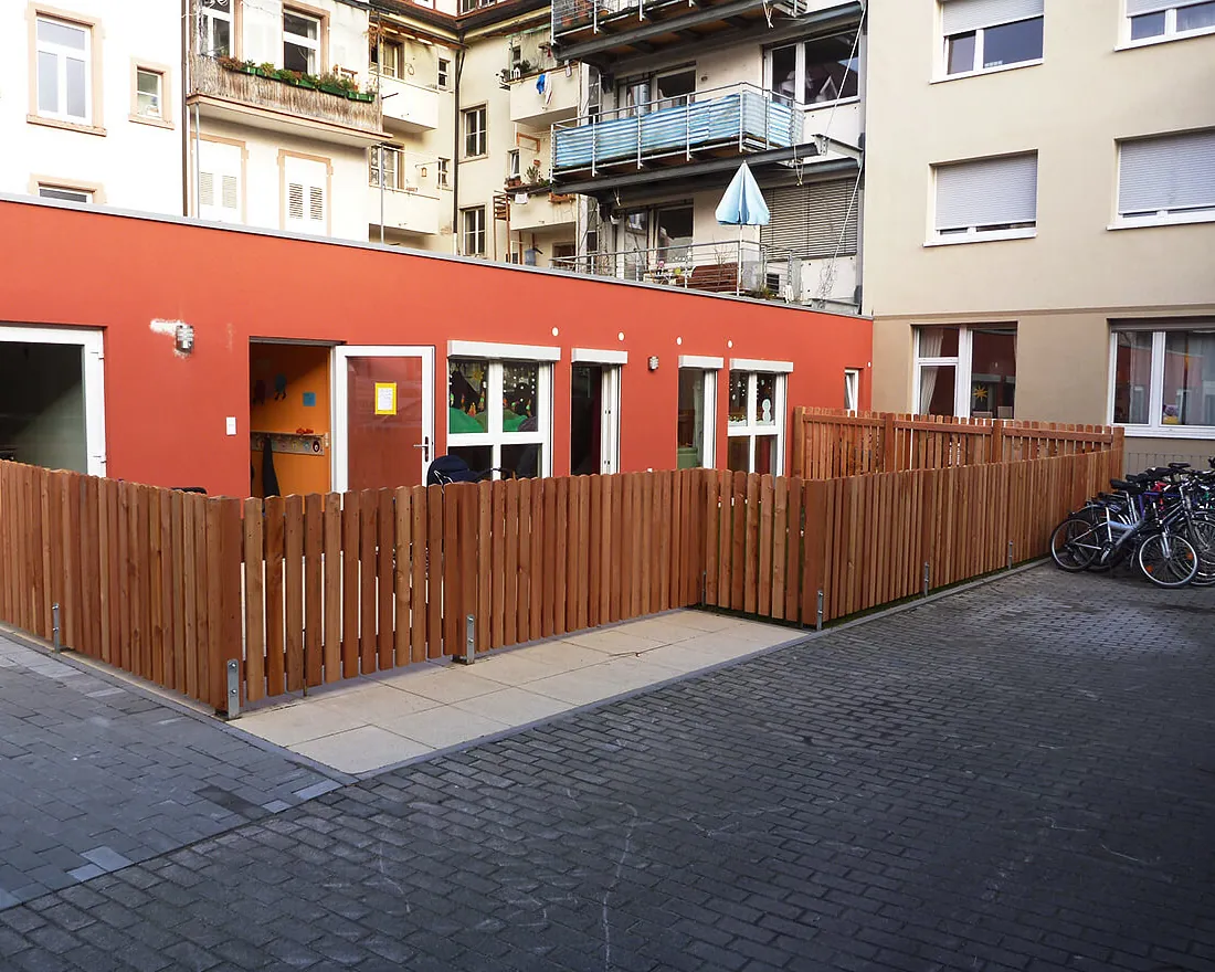 Außenbereich einer Kindertagesstätte mit roter Fassade, Holzzaun und mehreren Fenstern, umgeben von Wohngebäuden