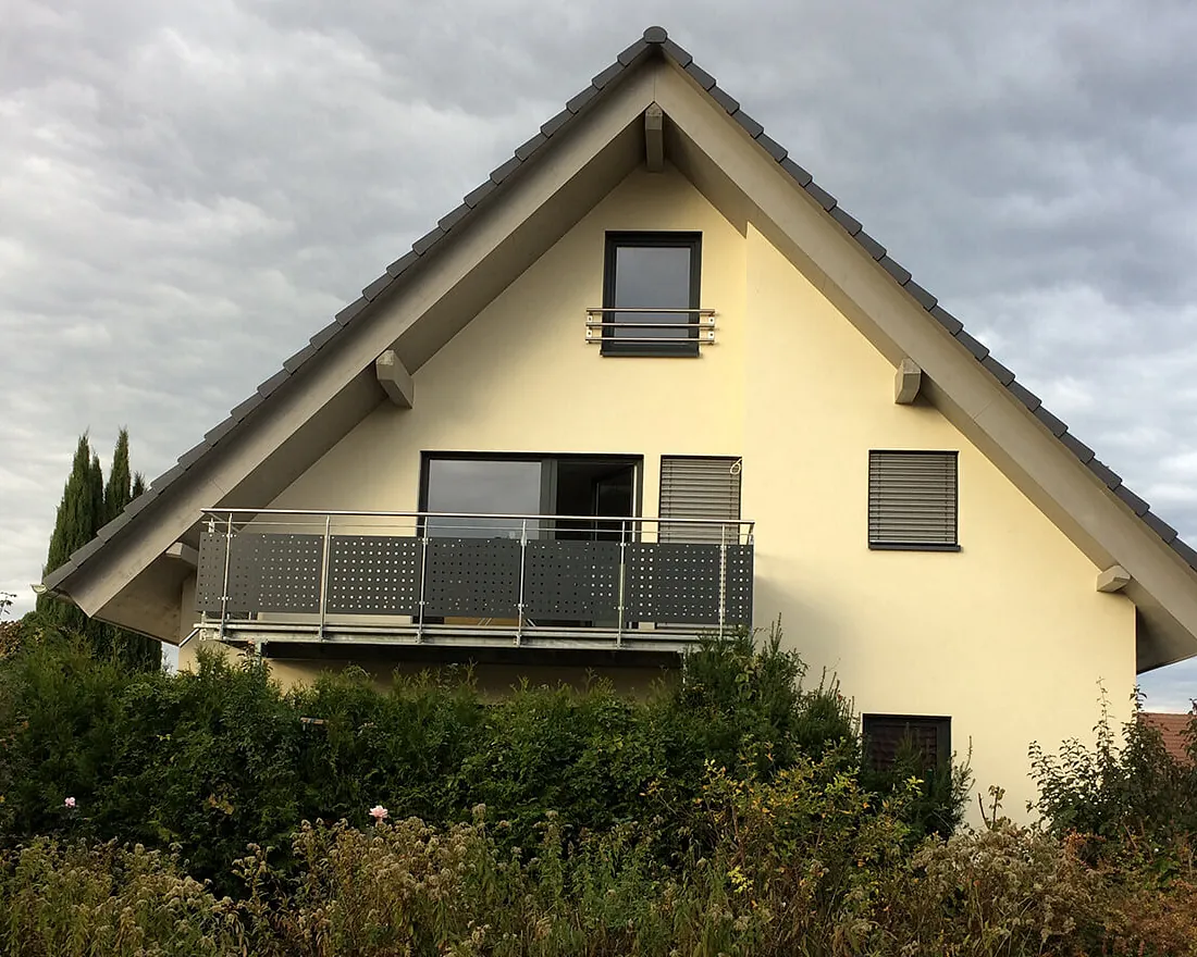 Zweistöckiges Einfamilienhaus mit gelber Fassade, großem Balkon und drei Fenstern unter bewölktem Himmel