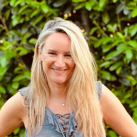 Frau mit langen, blonden Haaren, trägt ein ärmelloses, gestreiftes Oberteil und eine Halskette, vor grünem Laub