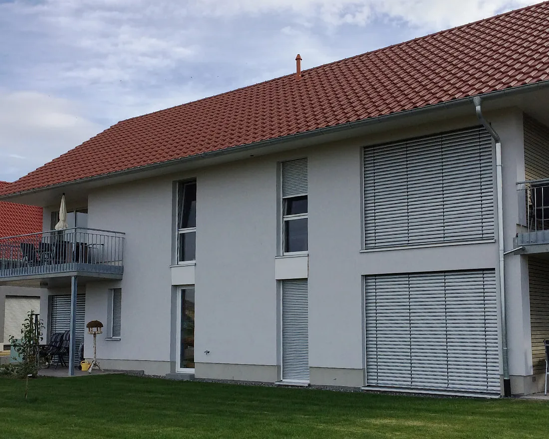 Zweistöckiges Ferienwohnhaus mit rotem Ziegeldach, weißen Wänden, mehreren Fenstern mit geschlossenen Jalousien und Balkon mit Sonnenschirm