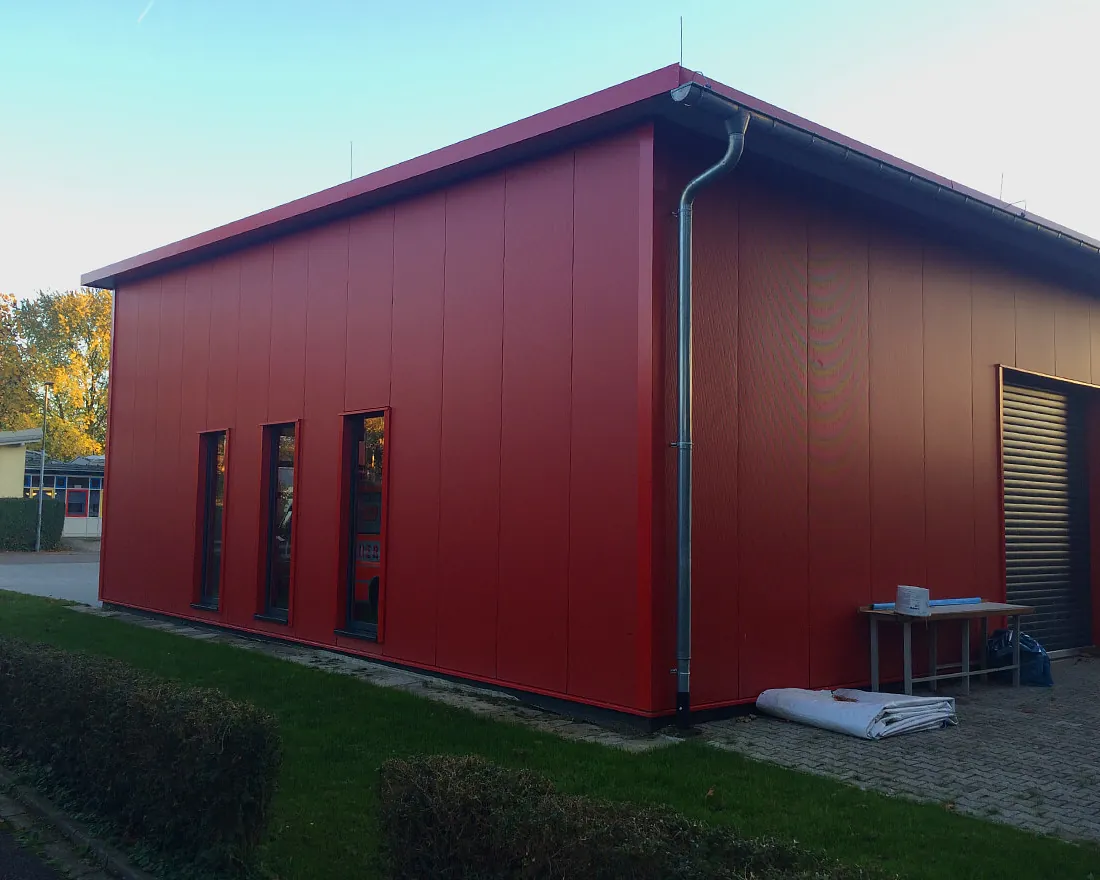 Rotes Feuerwehrgebäude mit vier schmalen Fenstern und einer großen Rolltoröffnung, umgeben von Grünfläche
