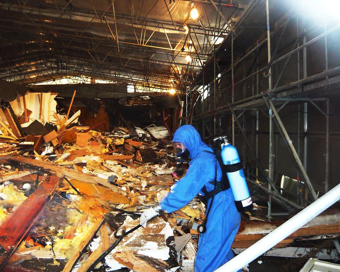 Person in blauem Schutzanzug und Atemgerät arbeitet in einer Halle mit Trümmern und Gerüststrukturen