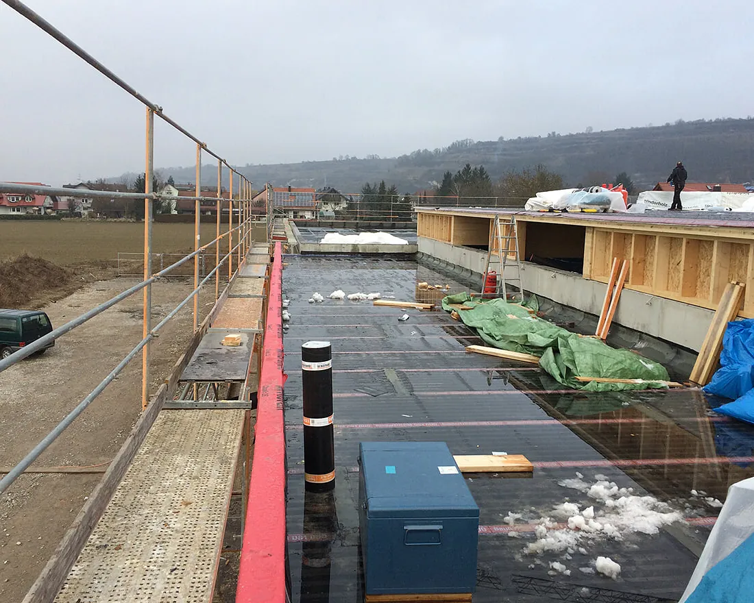 Baustelle eines Feuerwehrgerätehauses mit flachem Dach, auf dem Materialien und Bauausrüstung liegen, im Hintergrund Hügel und Häuser