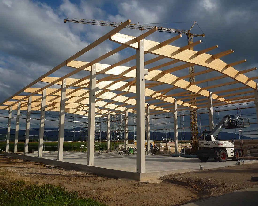 Rohbau einer großen Gewerbehalle mit Stahlbetonstützen und Holzbindern unter bewölktem Himmel